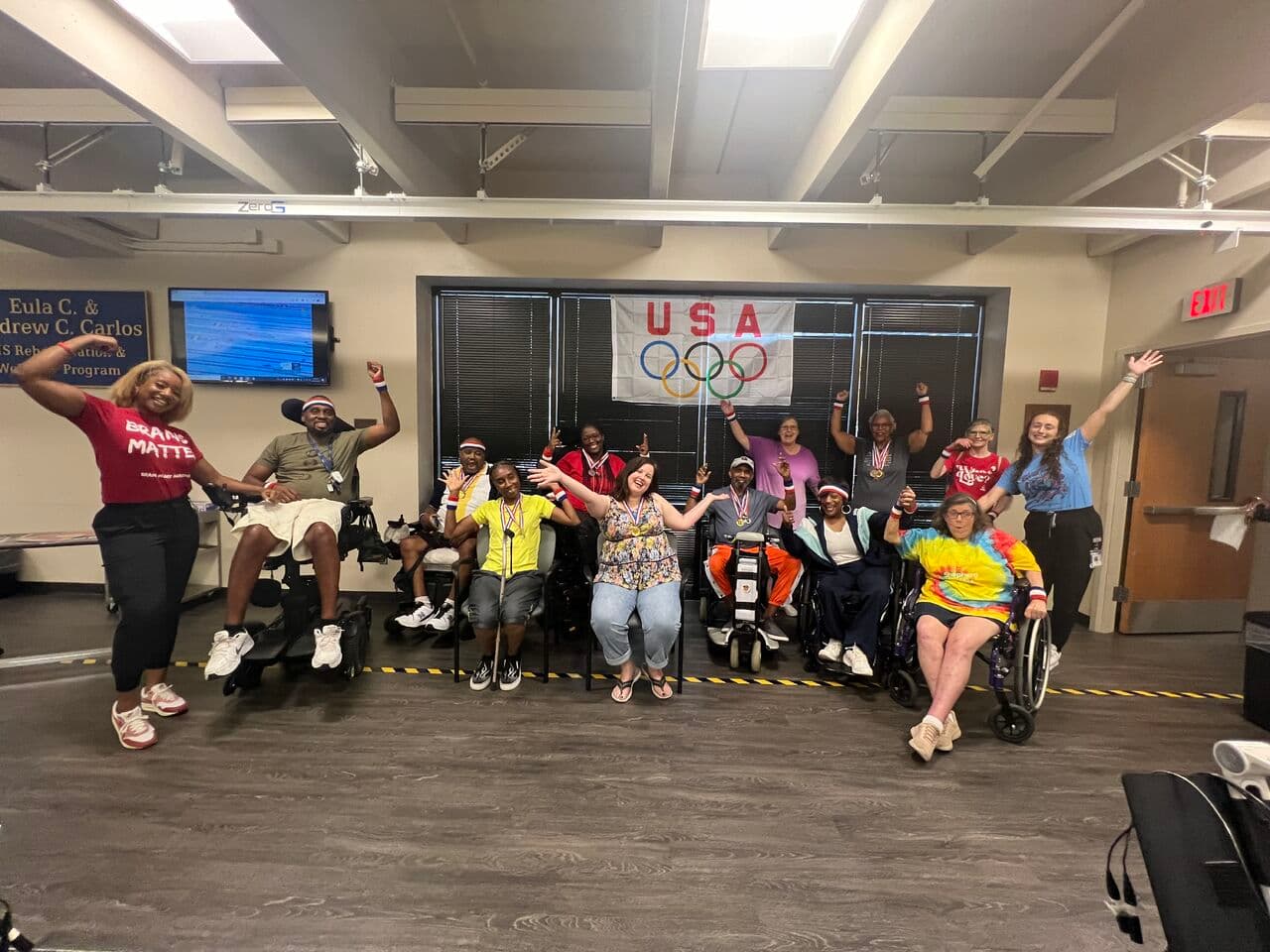 A diverse group of people, some in wheelchairs, are joyfully posing with arms raised in a room with a USA Olympics banner on the wall. They appear to be participating in a fitness or rehabilitation program.