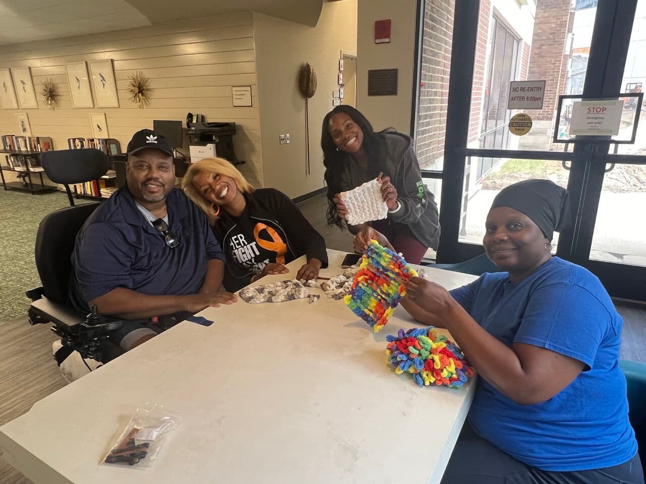 Four people sit around a table in a bright room, smiling at the camera while displaying handmade crafts. One person is seated in a wheelchair. The background shows a bookshelf and large windows.