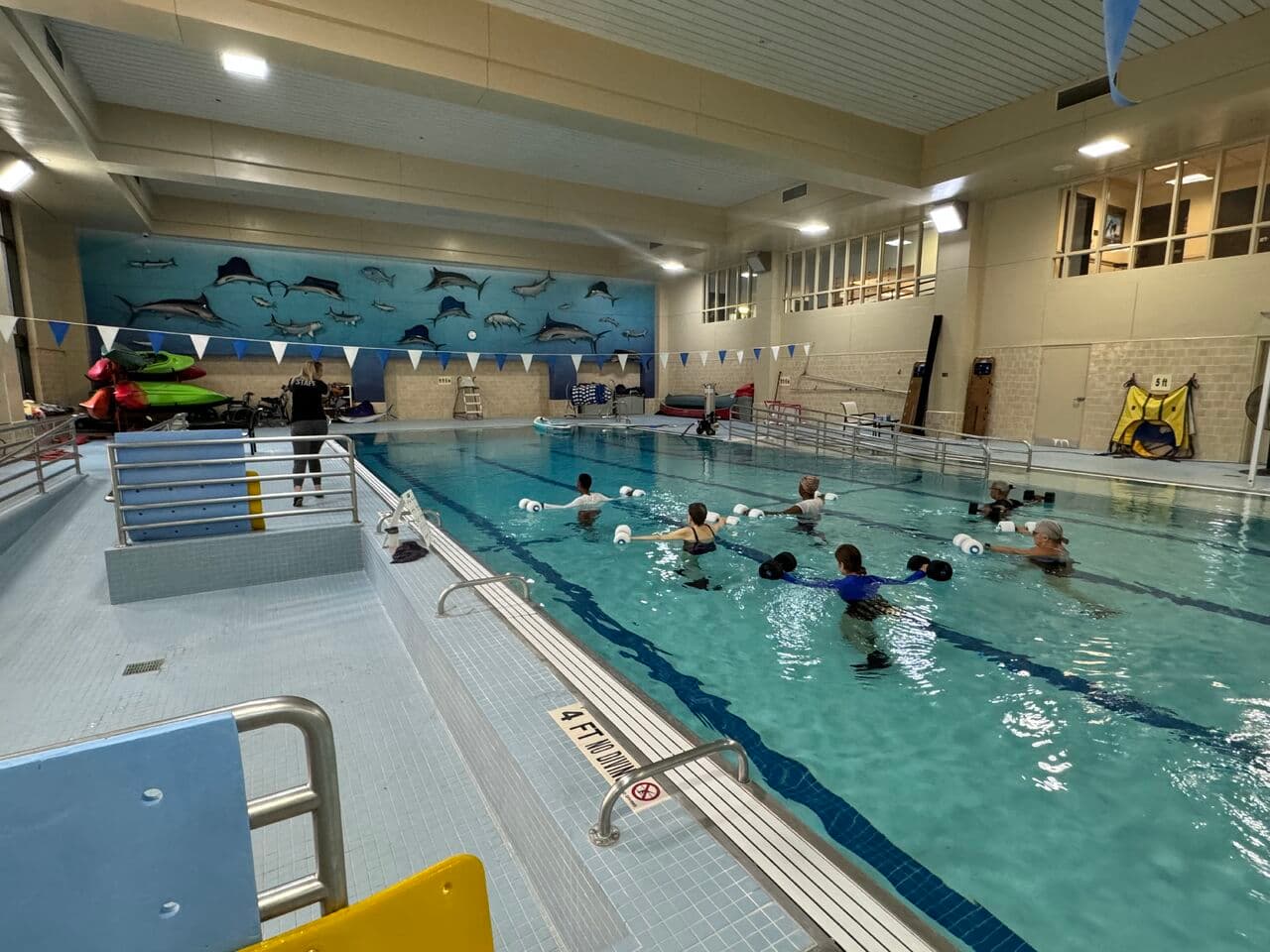 Indoor swimming pool with several people participating in a water exercise class. The pool is divided by lane markers, and a mural of marine life decorates the wall. The area is well-lit, and a few floatation devices are visible.