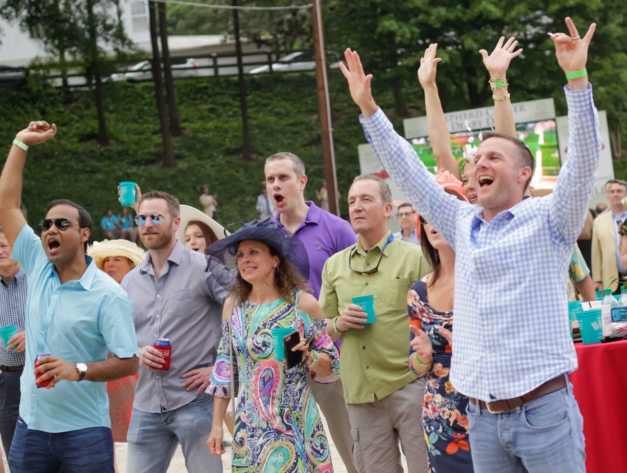 A group of people celebrating outdoors, raising their arms and holding drinks. They are dressed in casual, colorful attire, with some wearing hats. Trees and buildings can be seen in the background. The atmosphere is festive and joyful.