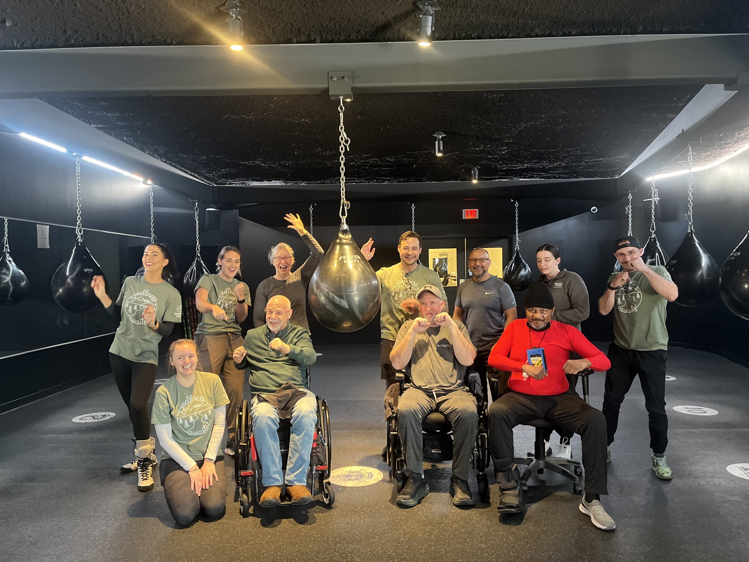 A group of diverse individuals, including two people in wheelchairs, pose happily in a gym with punching bags hanging behind them. They are all wearing casual workout clothes and smiling for the camera, exuding a sense of camaraderie and joy.