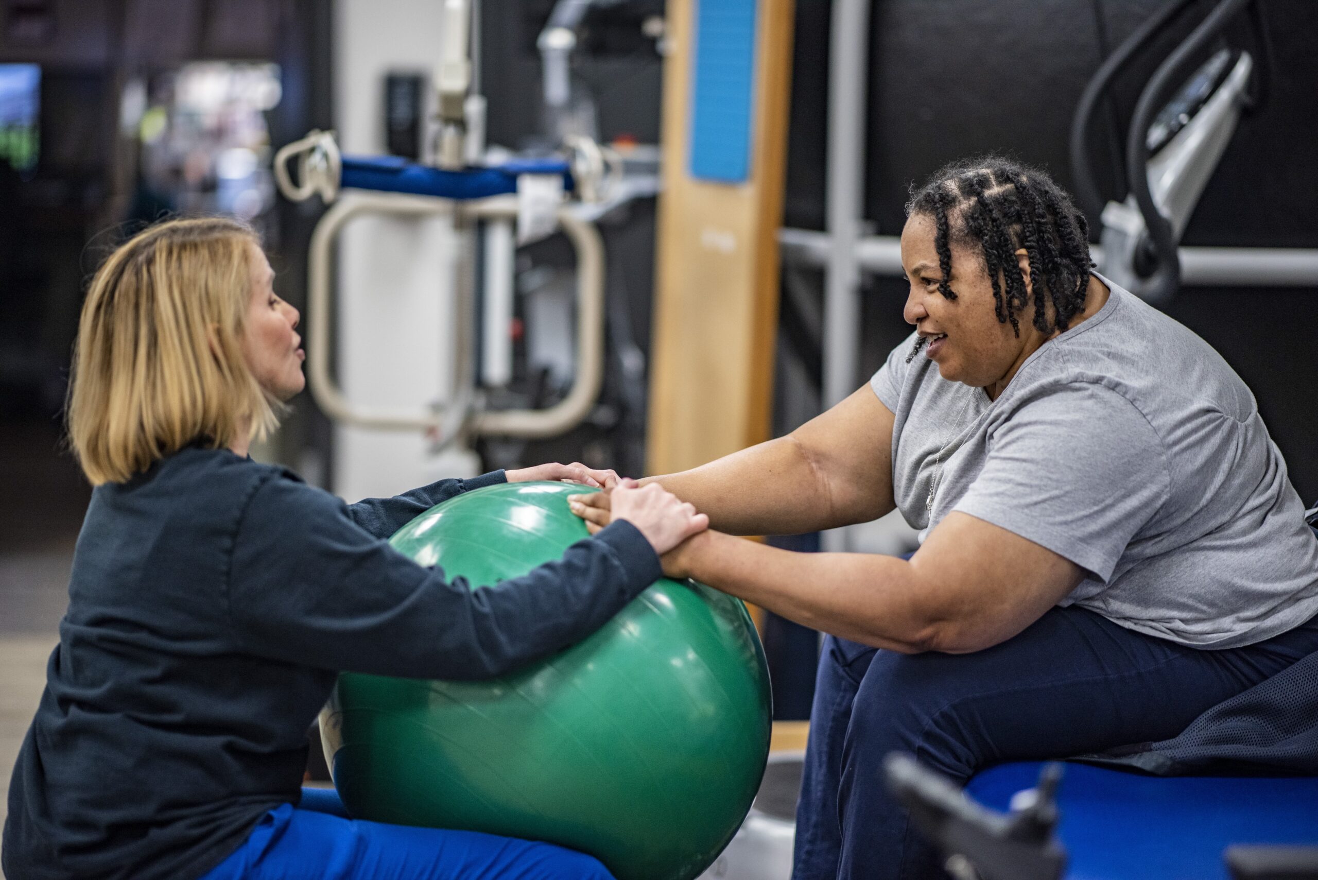 A healthcare worker assists a patient with rehabilitation exercises using a green exercise ball. They are seated facing each other, holding the ball between them in a fitness facility. Equipment and exercise machines are visible in the background.