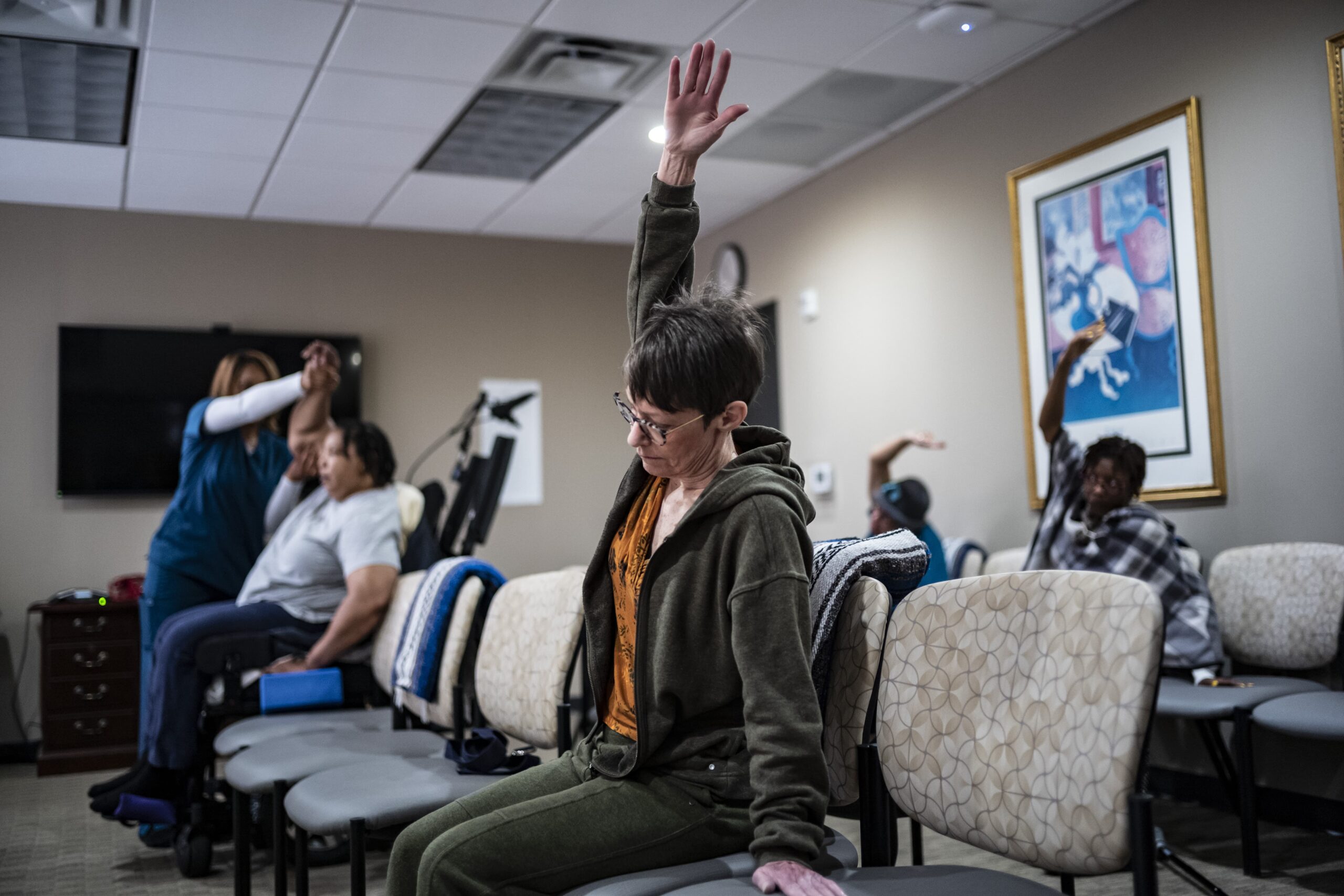 People sitting in a room are stretching with one arm raised. Some are in chairs, while a woman on the left assists another person. The setting appears to be a health or physical therapy space with a painting on the wall.