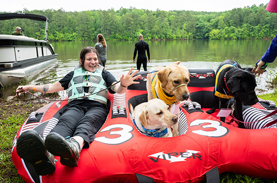 A woman in a life jacket sits on a red inflatable raft with two Labradors. The raft is near a lake shore with people and boats in the background. The scene is lively and outdoorsy, with trees surrounding the lake.