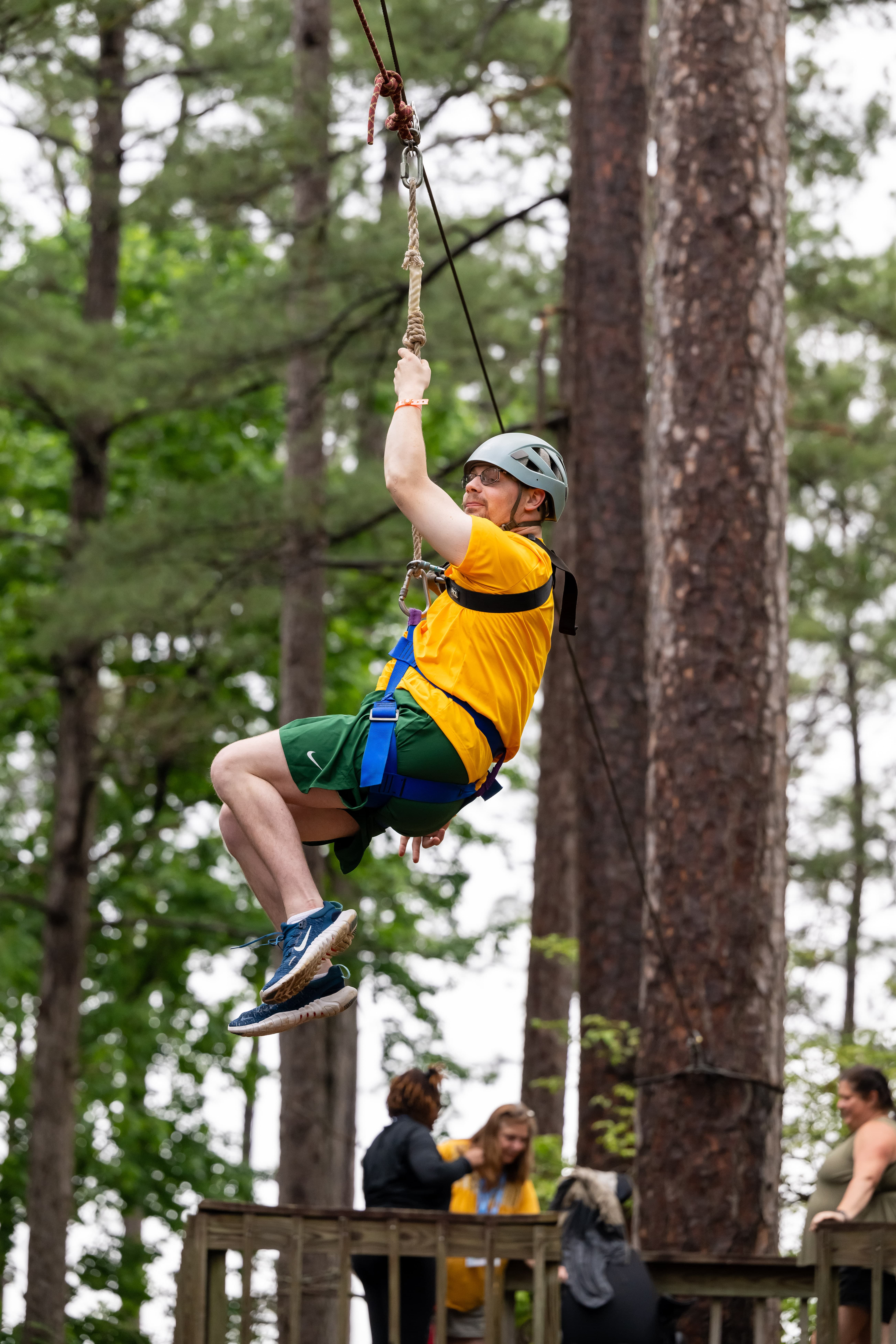 A person wearing a yellow shirt, green shorts, and a helmet is zip-lining through a forest area. The background shows tall trees and a group of people on a wooden platform watching.