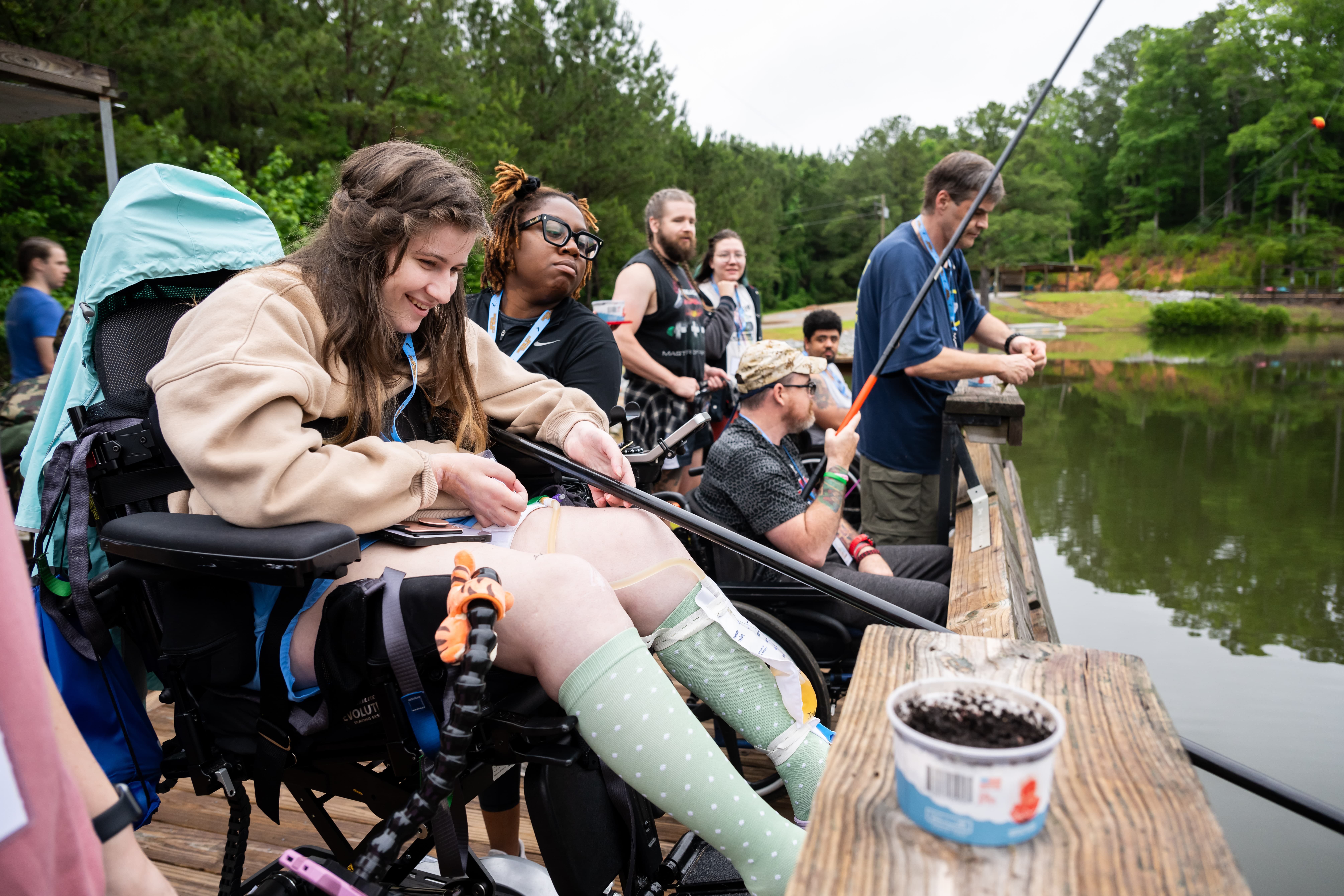 A group of people, including a woman in a wheelchair, are fishing by a lakeside. The scene is set on a wooden deck with trees in the background. They appear engaged and enjoying the activity together, with fishing gear and a bait container visible.