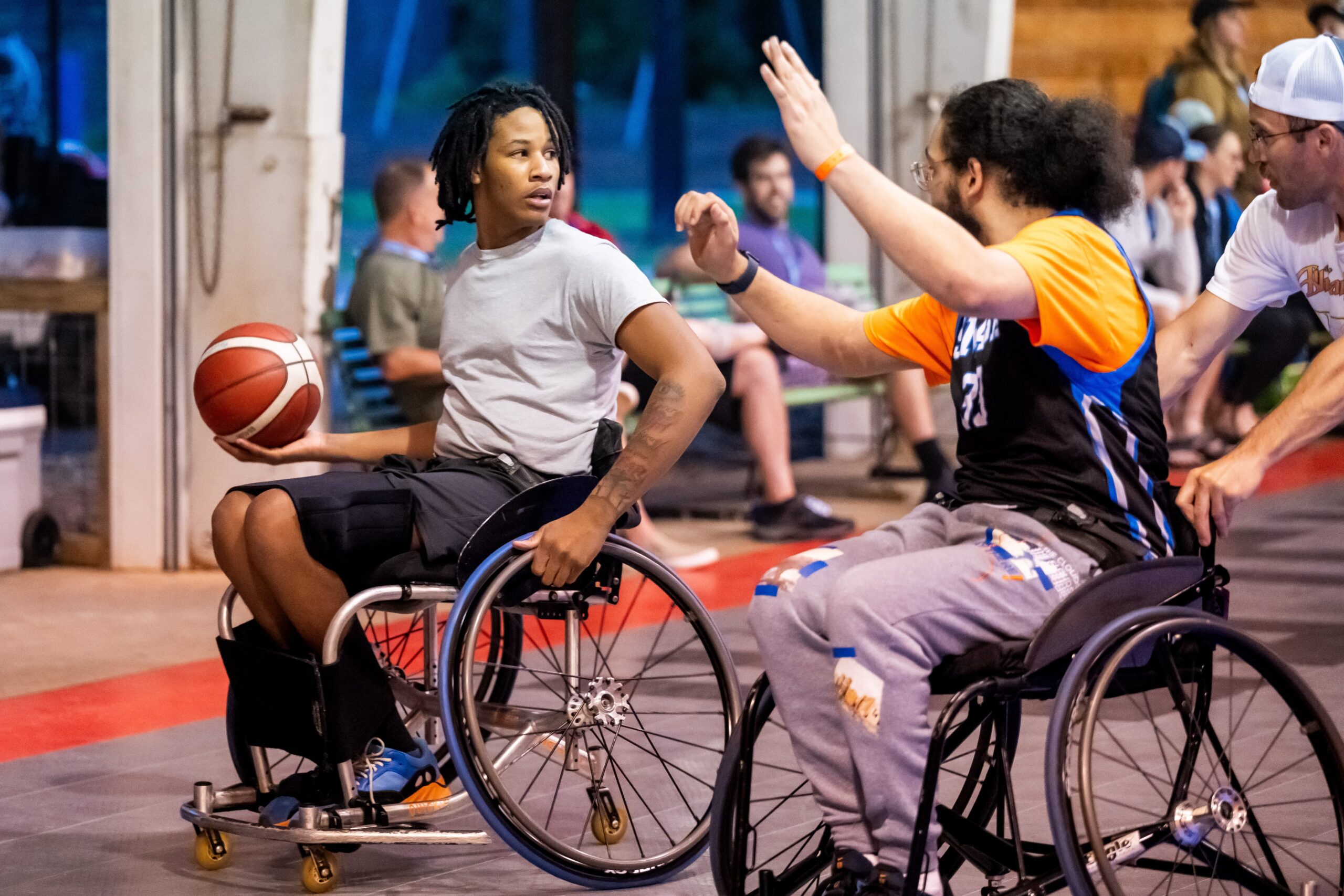 Two men in wheelchairs play basketball indoors. One, in a gray shirt, holds the ball and looks determined. The other, in a blue and orange jersey, raises his arm to block. People watch in the background.