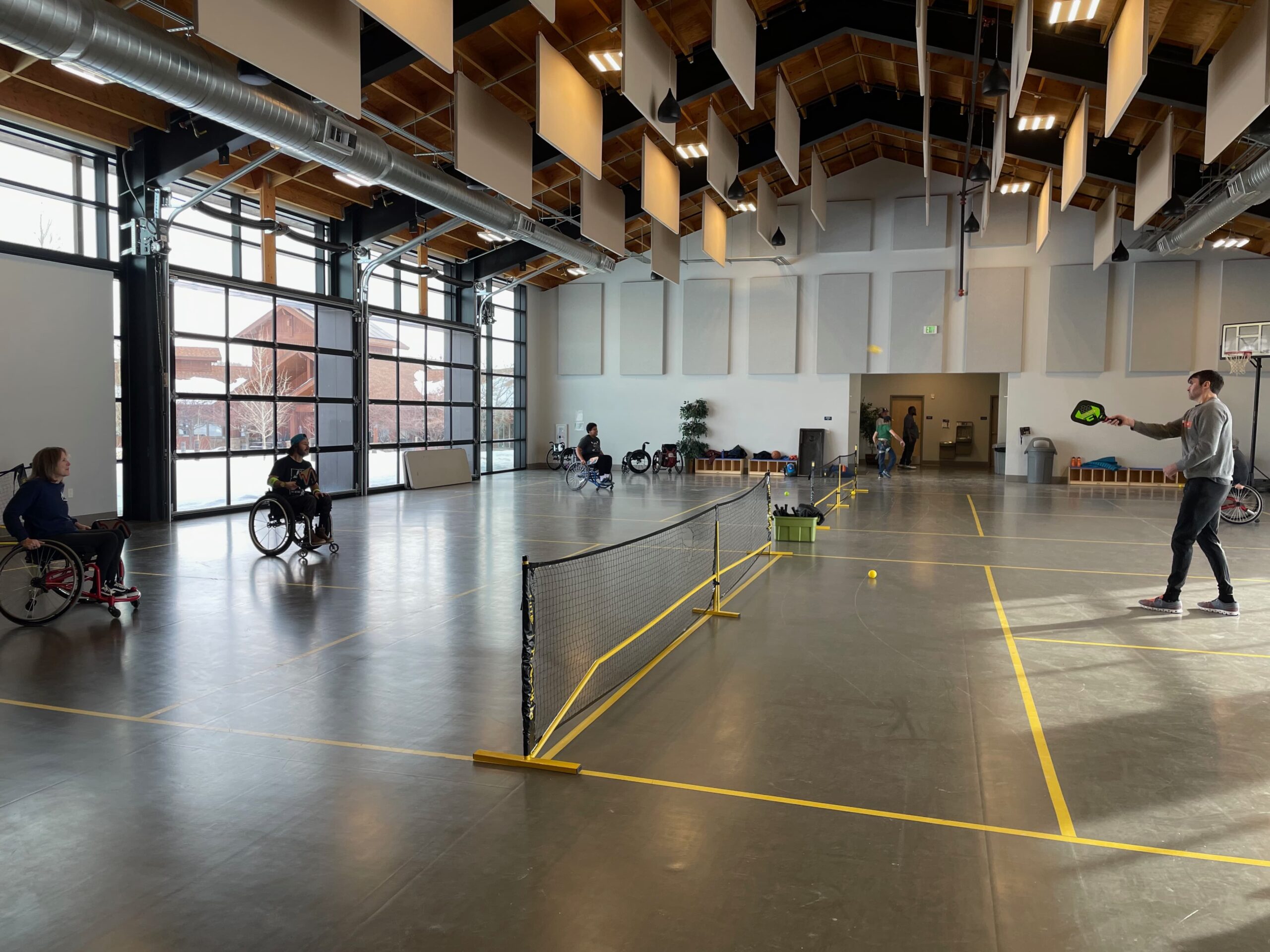 A spacious indoor tennis court with a high ceiling. Three people in wheelchairs on one side of a net, and a person standing on the other side, hitting a tennis ball. Sunlight streams in through large windows.