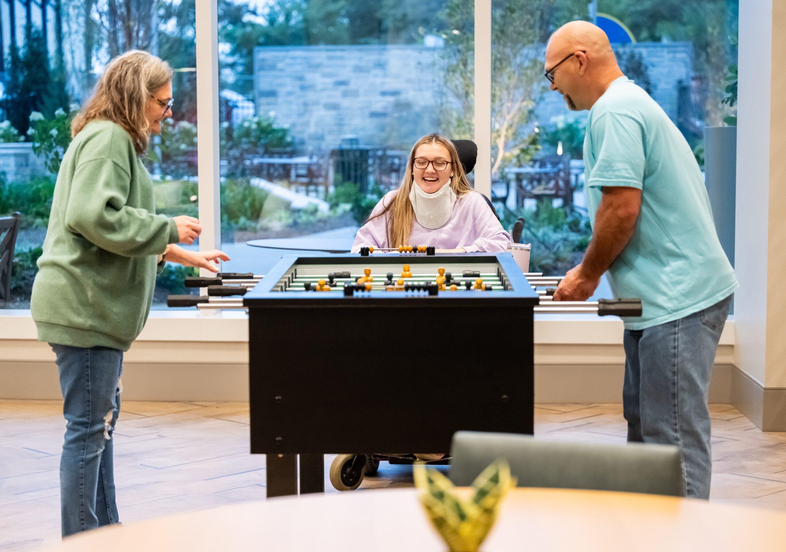 Three people are playing a game of foosball indoors. A woman with long hair is sitting in a wheelchair, smiling. Two other individuals stand on either side of the table, actively engaged in the game. Large windows are in the background.