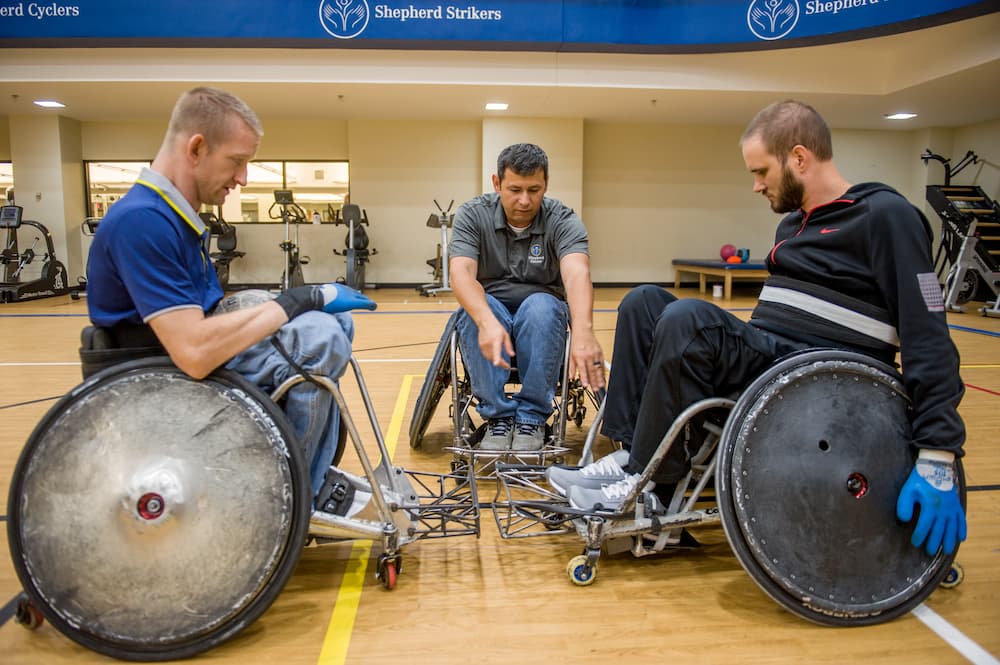 Three people in wheelchairs are on a basketball court. Two are wearing gloves and facing each other with their wheels angled slightly. A third person in the middle appears to be instructing them. Gym equipment is in the background.