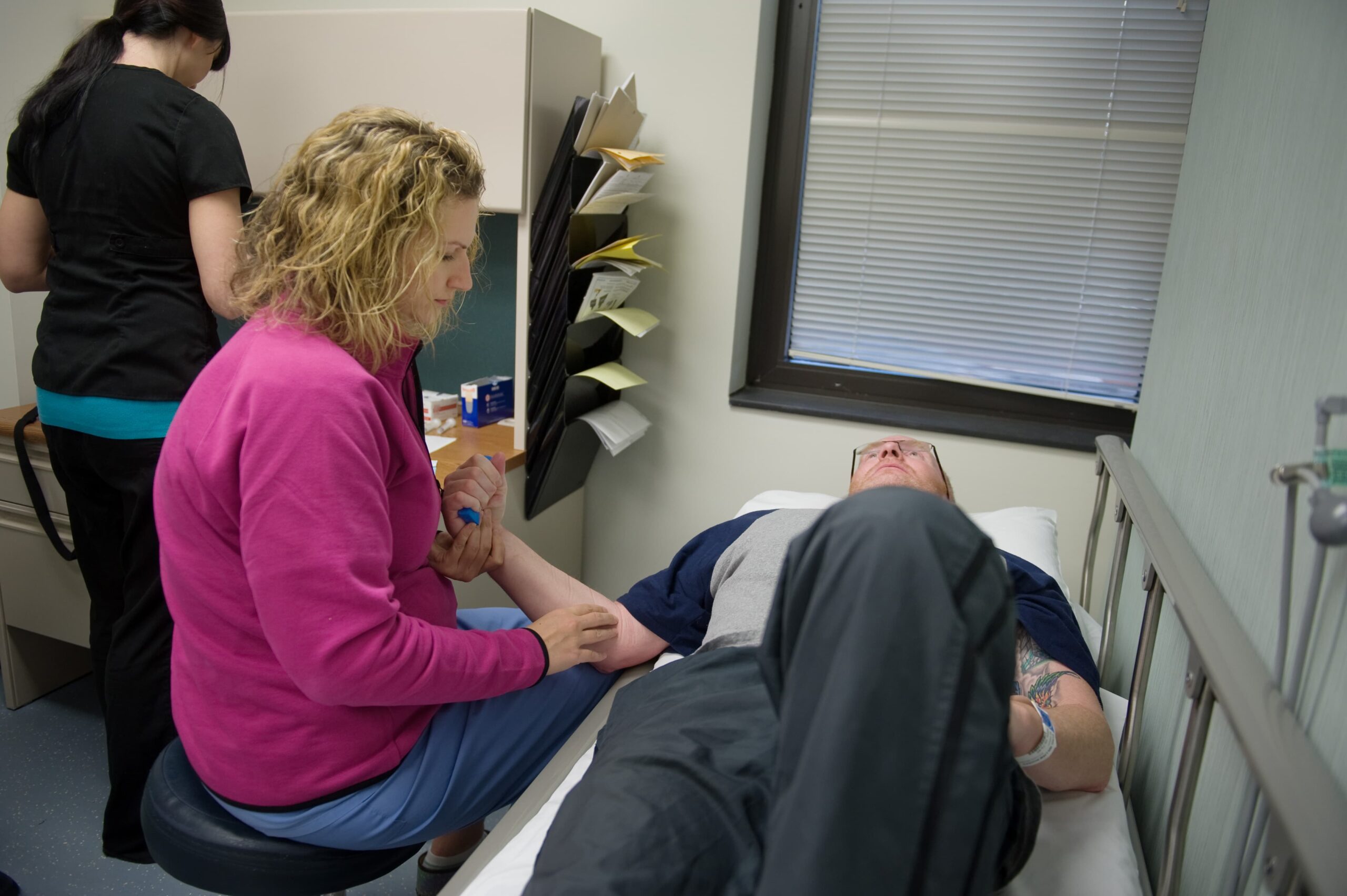 A medical professional in a pink jacket is drawing blood from a patient lying on a bed. The patient is wearing a gray and blue shirt. Another person in black scrubs is standing at a counter in the background. A window with blinds is on the wall.