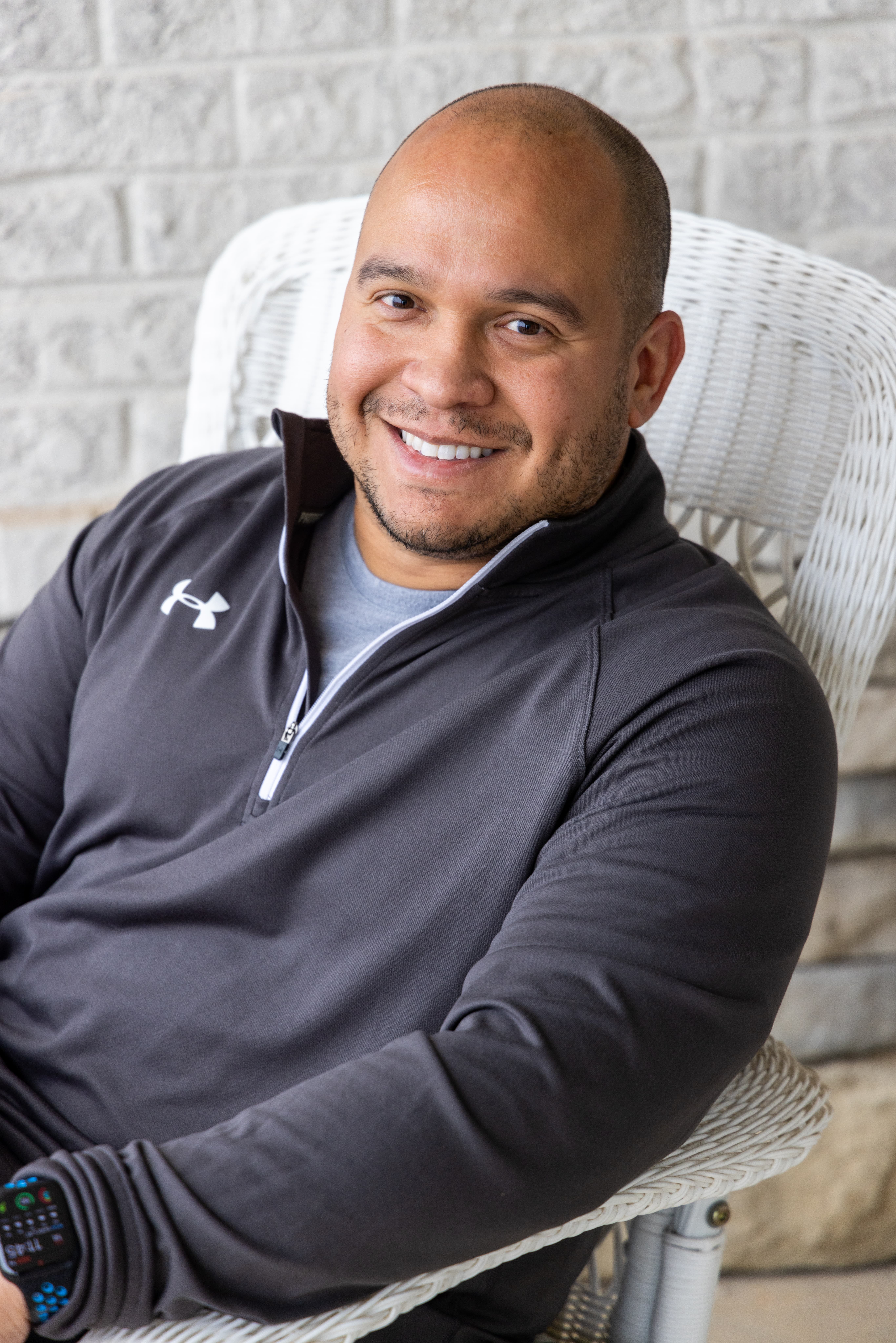 Smiling man in a gray Under Armour zip-up jacket sits in a white wicker chair against a light brick wall. He has a smartwatch on his wrist and short hair.