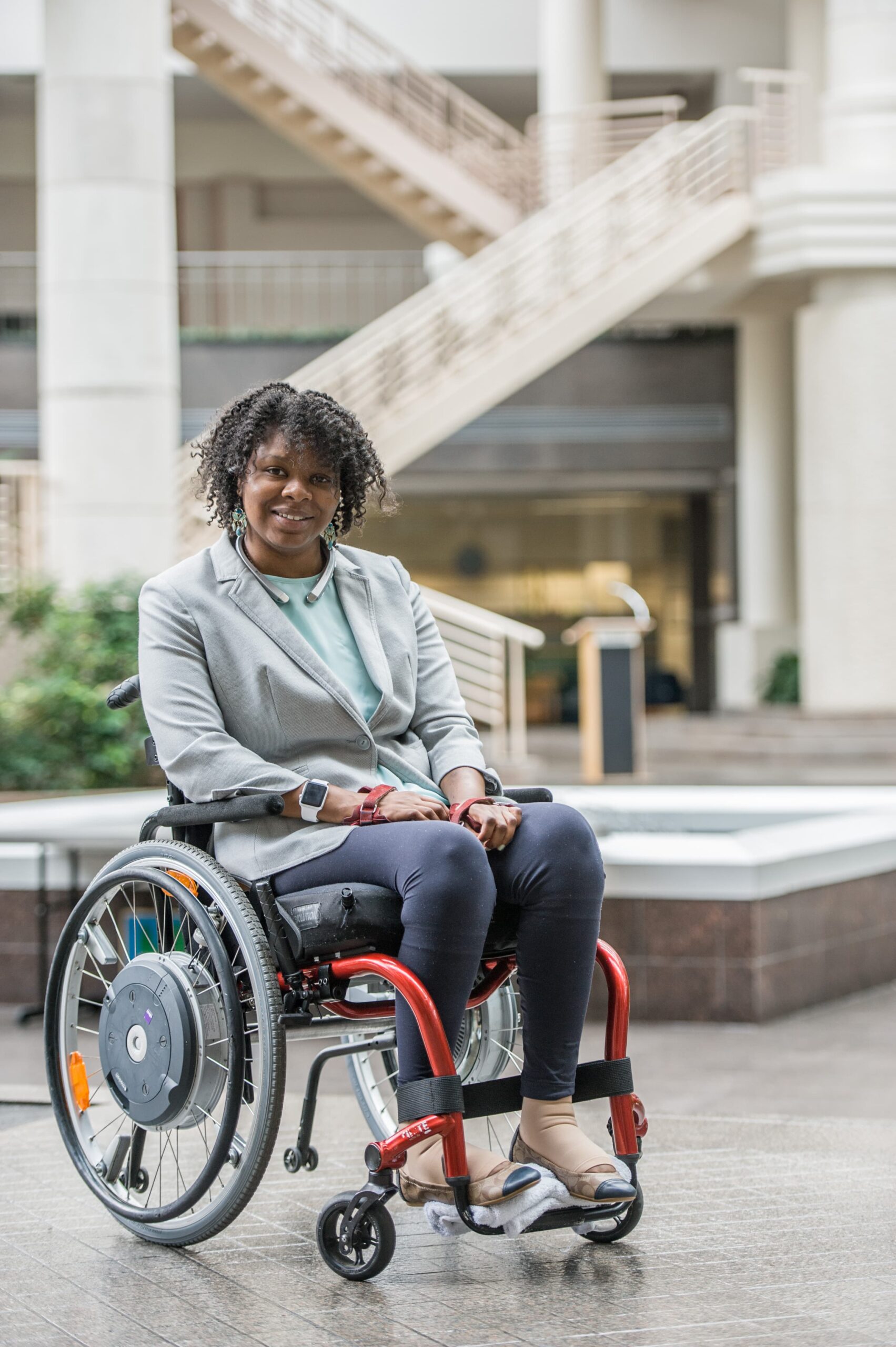 A woman sitting in a wheelchair, smiling, wearing a gray blazer and dark pants. She is situated in a spacious indoor area with a staircase and greenery in the background.