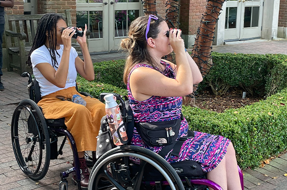 Two people in wheelchairs, one wearing a white shirt and orange skirt, and the other in a purple patterned dress, use binoculars outdoors. They're in front of a brick building with greenery and a tree wrapped in string lights nearby.