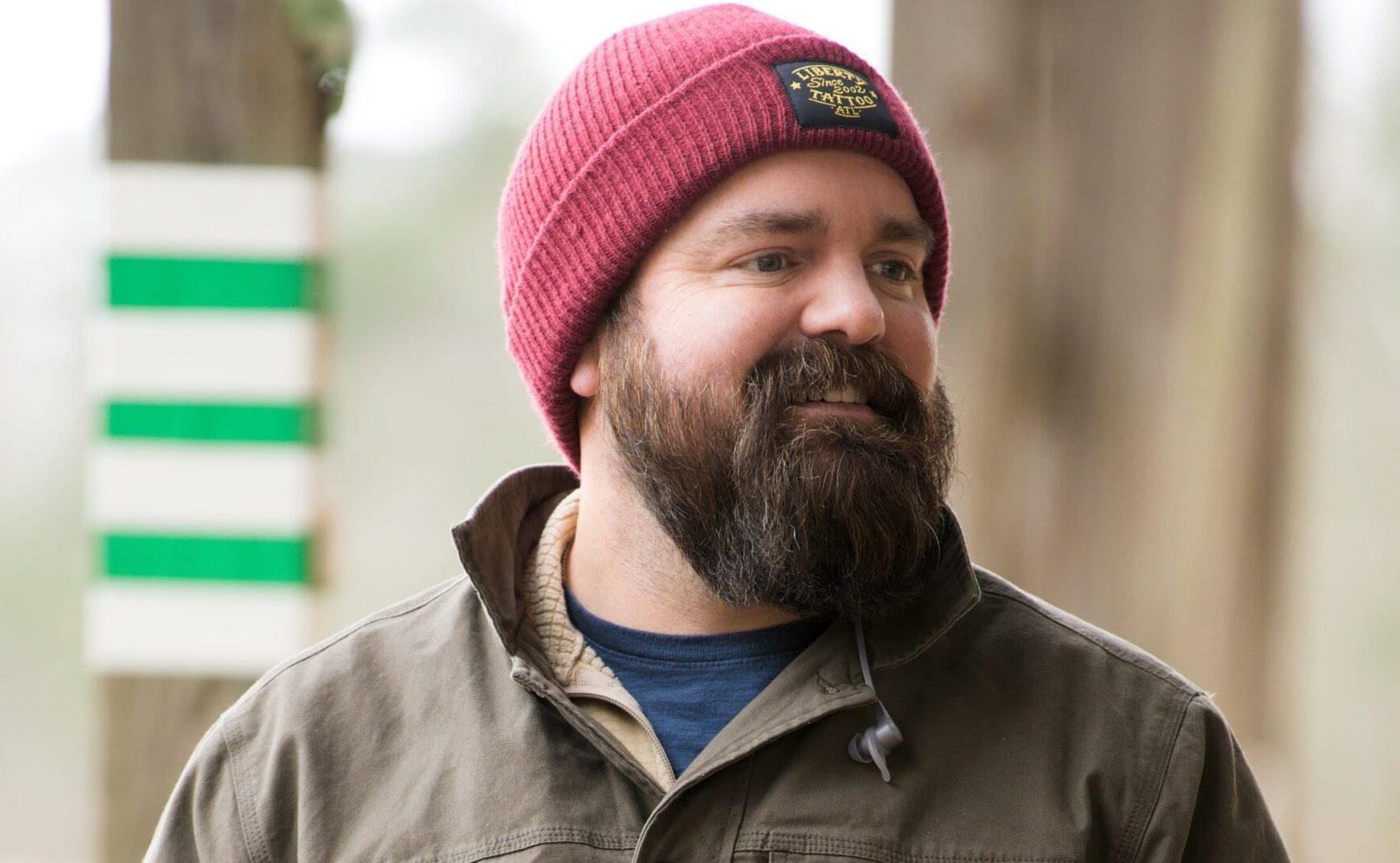 A person with a beard wears a red beanie and a green jacket while standing outdoors. They appear relaxed and are looking to one side with a slight smile. The background is blurred with natural elements visible.