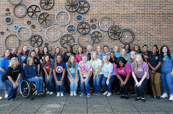 A group of people wearing matching blue T-shirts pose together in front of a brick wall decorated with various bicycle wheels.