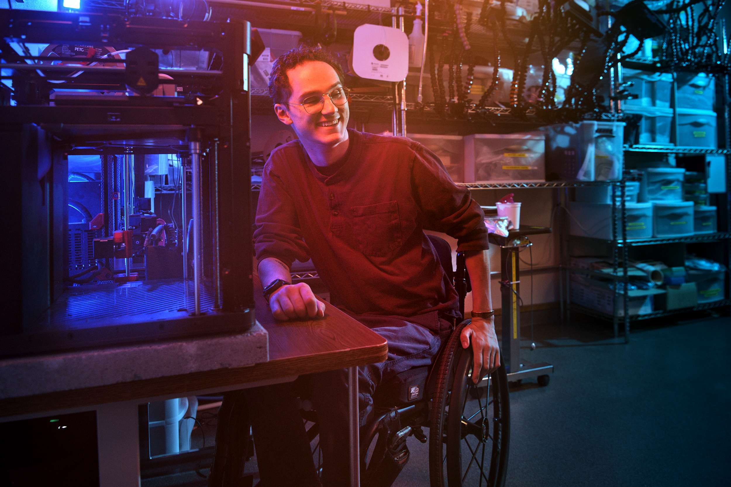 A person in a wheelchair smiles while sitting near a 3D printer in a workshop. The room is filled with shelves and equipment, illuminated by blue and red lights, creating a futuristic ambiance.