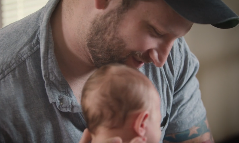 A bearded man in a cap, wearing a gray shirt, lovingly holds and looks at a baby. The baby is wearing a light blue outfit, resting against the man's shoulder. The image captures a tender moment between them.