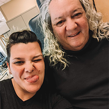 Two people smiling at the camera. The person on the left is making a playful expression with their tongue out, and the person on the right has long curly hair. Both are wearing dark shirts. They appear to be in a casual indoor setting.