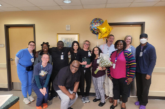 A group of twelve hospital staff members pose together in a hallway. They are smiling, with one person holding balloons and another holding a bouquet. Some are in scrubs, and one person is wearing a mask.