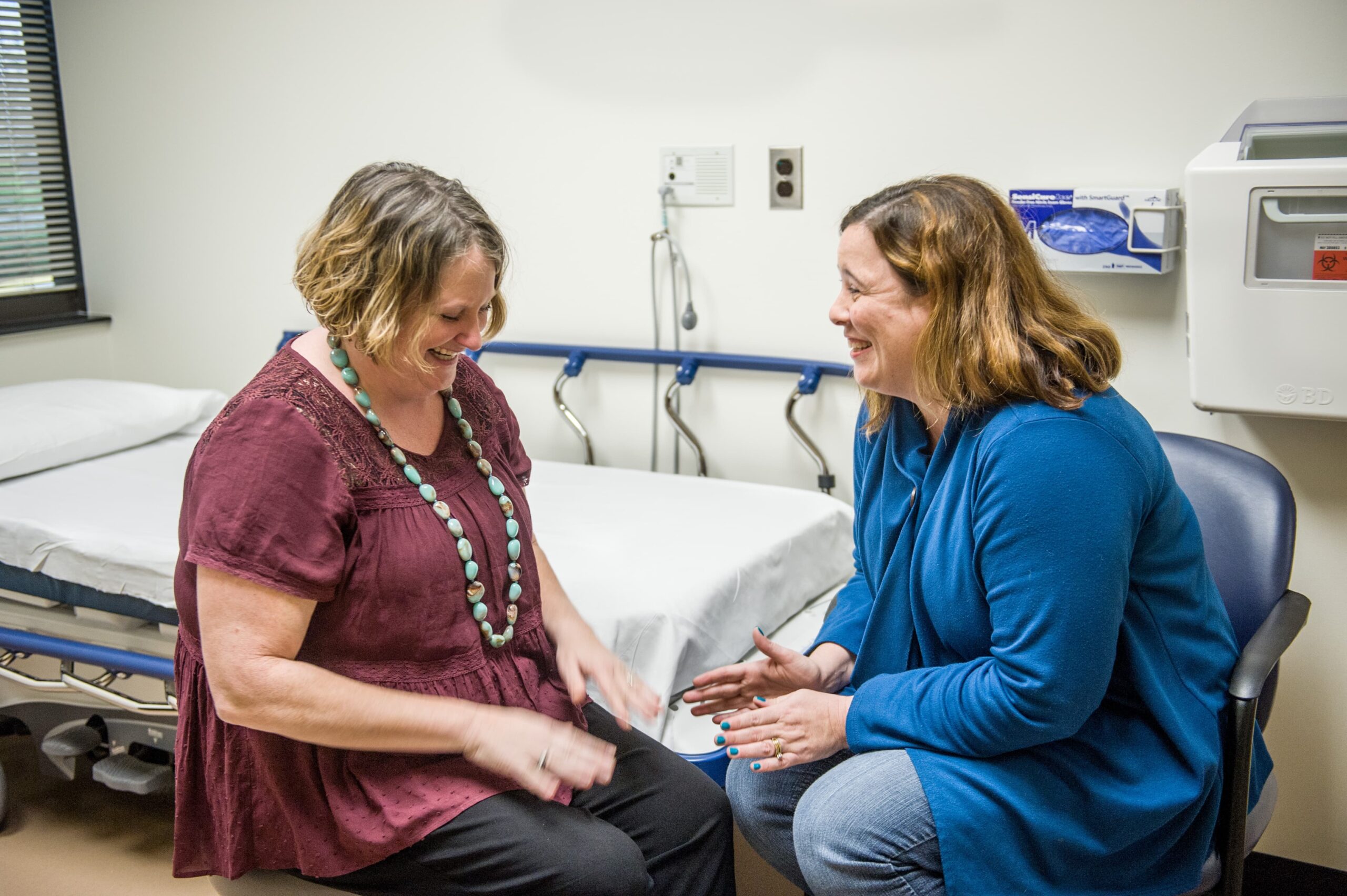 Two women sitting and smiling in a medical examination room. One is wearing a maroon blouse and necklace, the other a blue cardigan. They are engaged in conversation, with an exam table and medical equipment visible in the background.