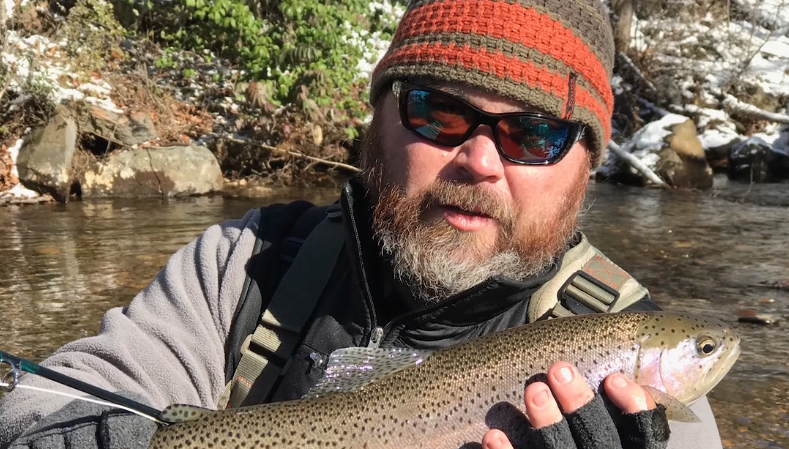 A person wearing a knit cap and sunglasses holds a large fish, possibly a trout, near a stream. The individual is dressed in outdoor gear, and the background includes rocks and greenery with some snow.