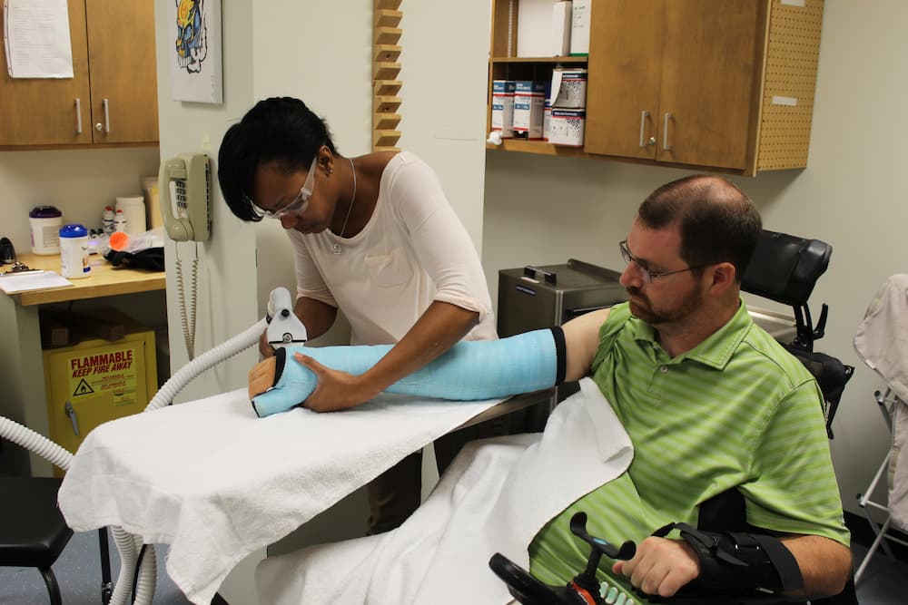 A healthcare professional assists a man in a green striped shirt and glasses with his blue cast on his arm at the renowned Shepherd Center. The man sits in a wheelchair, receiving upper extremity treatment, with a white towel covering part of his lap and arm. Shelves and medical equipment are in the background.
