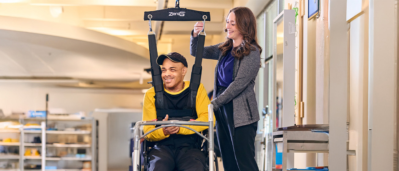 A man in a harness is supported by two women during a rehabilitation session. One woman adjusts the harness, while the other holds the man's hand. They are in a well-lit indoor room, all smiling and engaging positively.