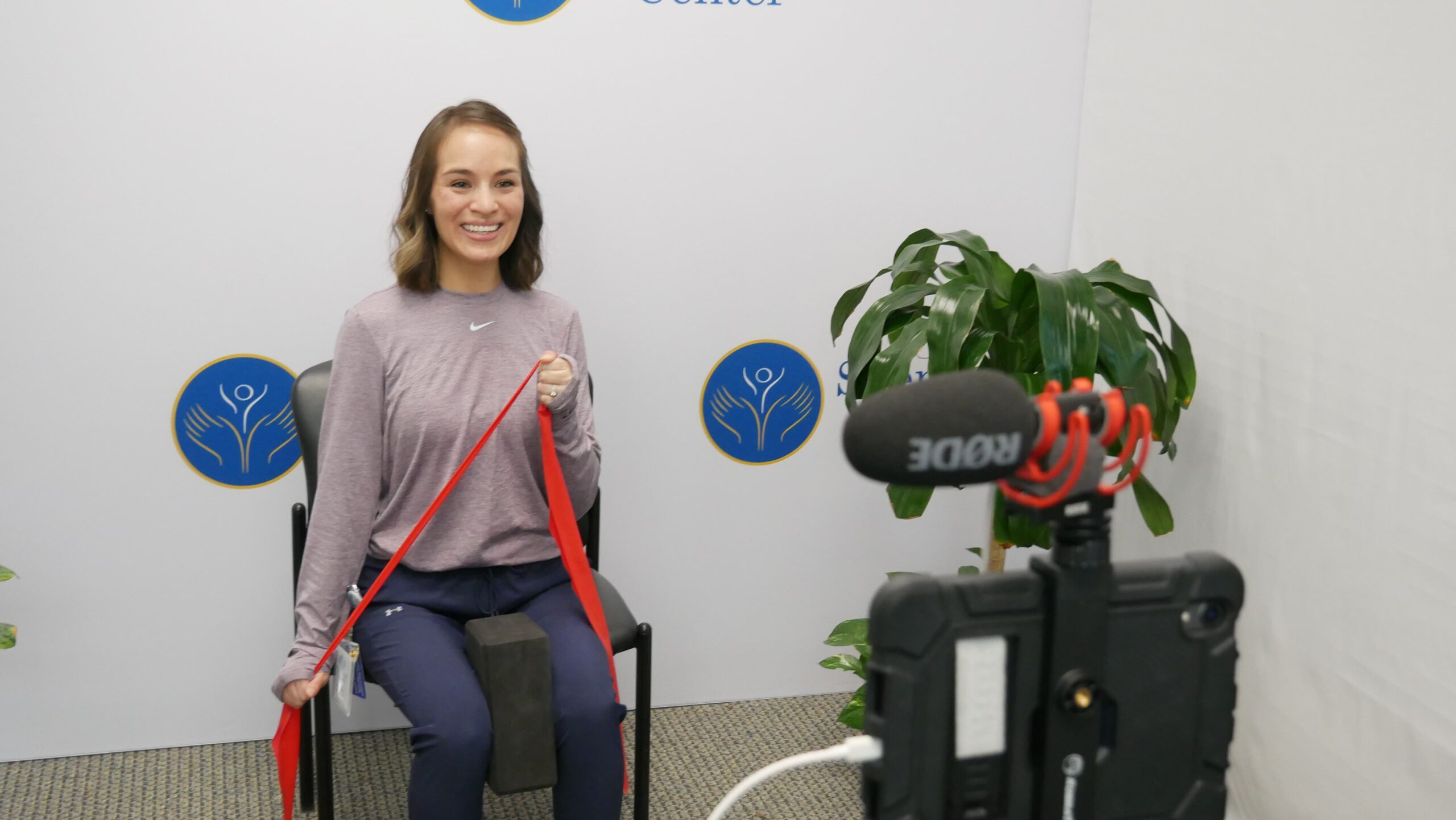 A woman in workout attire sits on a chair, smiling at the camera. She holds a red resistance band. A microphone on a stand is in the foreground, and there are potted plants in the background.