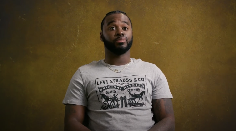 A person with a beard and braided hair is sitting against a brown textured background. They are wearing a gray Levi Strauss & Co. T-shirt with a graphic design and a necklace. The person has a neutral expression and their hands are resting on their lap.