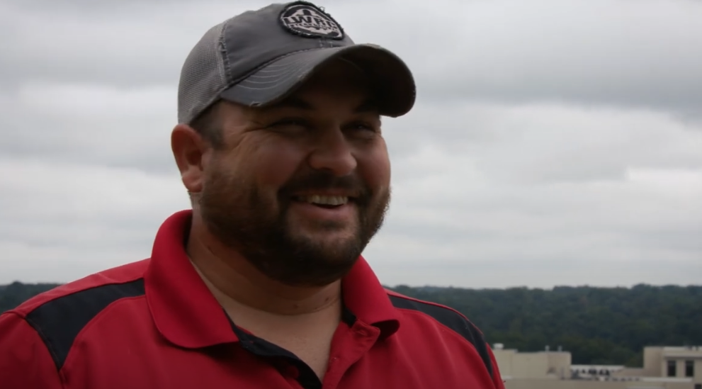 A man with a beard wearing a gray cap and a red and black collared shirt is smiling outdoors. In the background, there are trees and a cloudy sky.