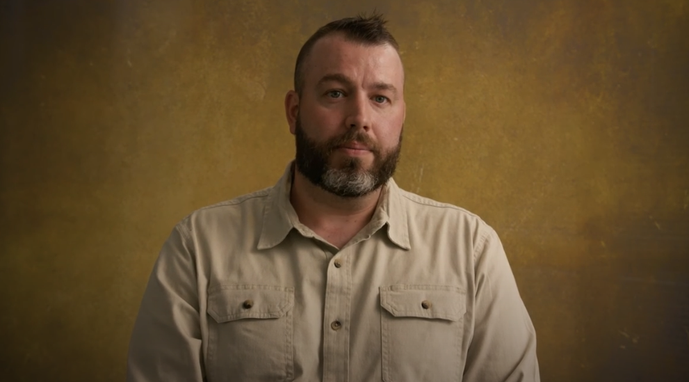 Man with a beard and a short haircut wearing a beige button-up shirt sits against a textured yellow-brown background, looking directly at the camera.