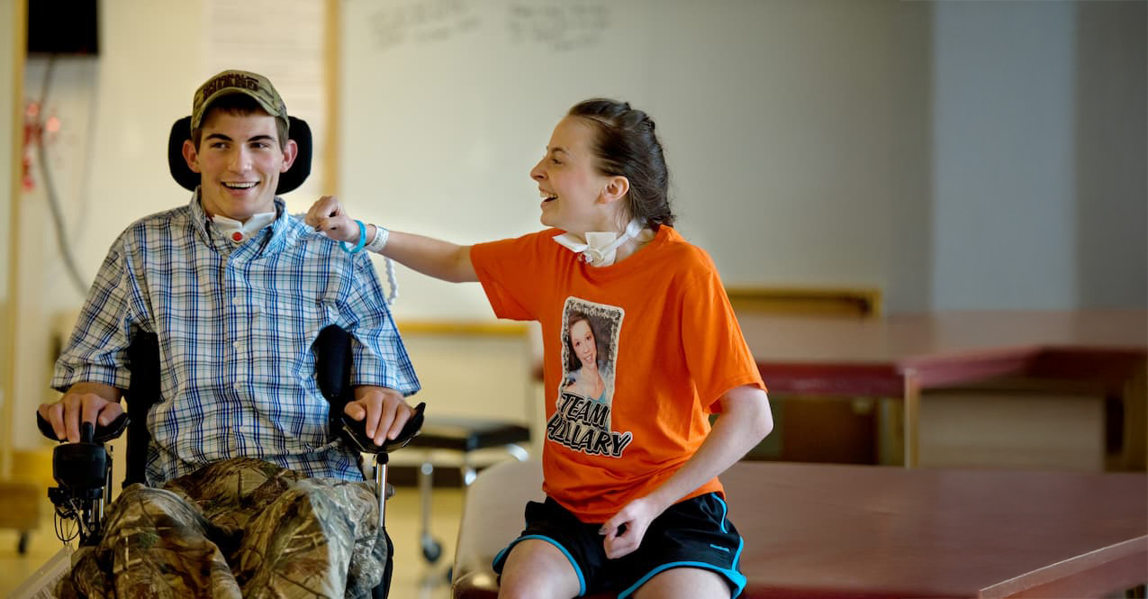 In a brightly lit room, a young man in a wheelchair, dressed in a plaid shirt and camouflage pants, smiles as he's playfully touched by a woman in an orange 