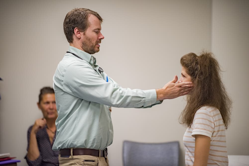 A man in a green shirt examines a woman's face in a clinical setting. The woman, wearing a white and gray striped shirt, stands facing him. Another person seated in the background is watching the interaction.