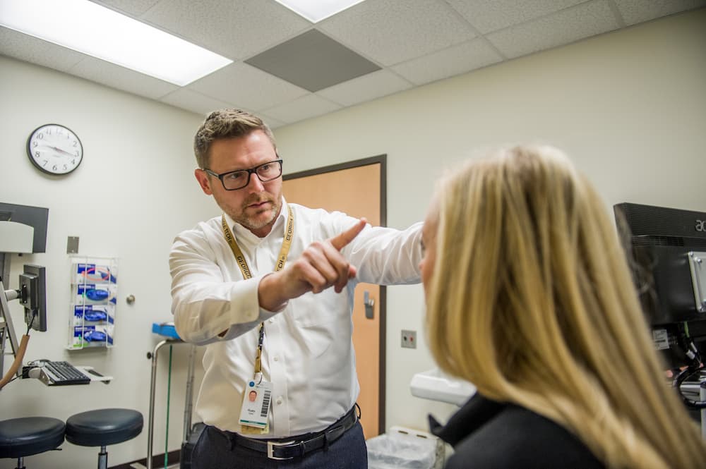 A healthcare professional is examining a patient in a medical office. He is pointing upwards while the patient, with long blonde hair, sits facing him. Medical equipment is visible in the background.