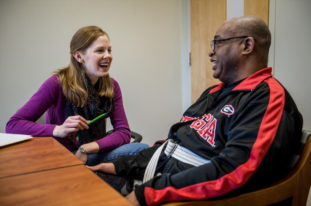 A woman with long hair sits at a table, smiling and holding a pen, facing a man in a Georgia jacket who is seated in a wheelchair. Both appear to be engaged in a friendly conversation.