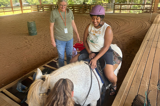 A person wearing a purple helmet smiles while sitting on a white horse inside an indoor arena. Two women stand nearby, one with a lanyard and the other adjusting the saddle. Natural light filters in, illuminating the sandy floor and green trees outside.