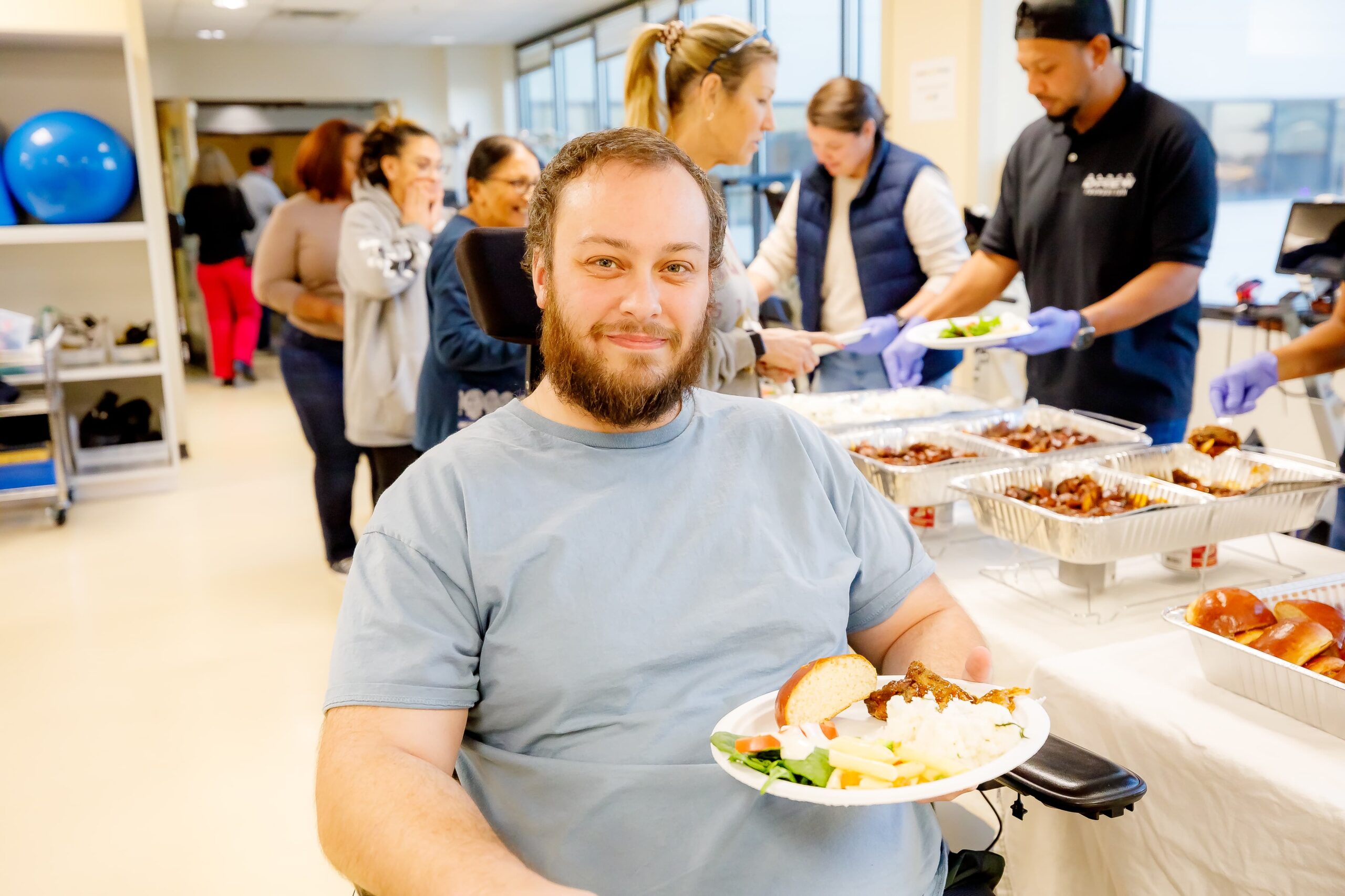 A man in a blue shirt, sitting in a wheelchair, smiles while holding a plate of food. Behind him, several people are serving and picking up food from a buffet table. The setting appears to be a community event or gathering.