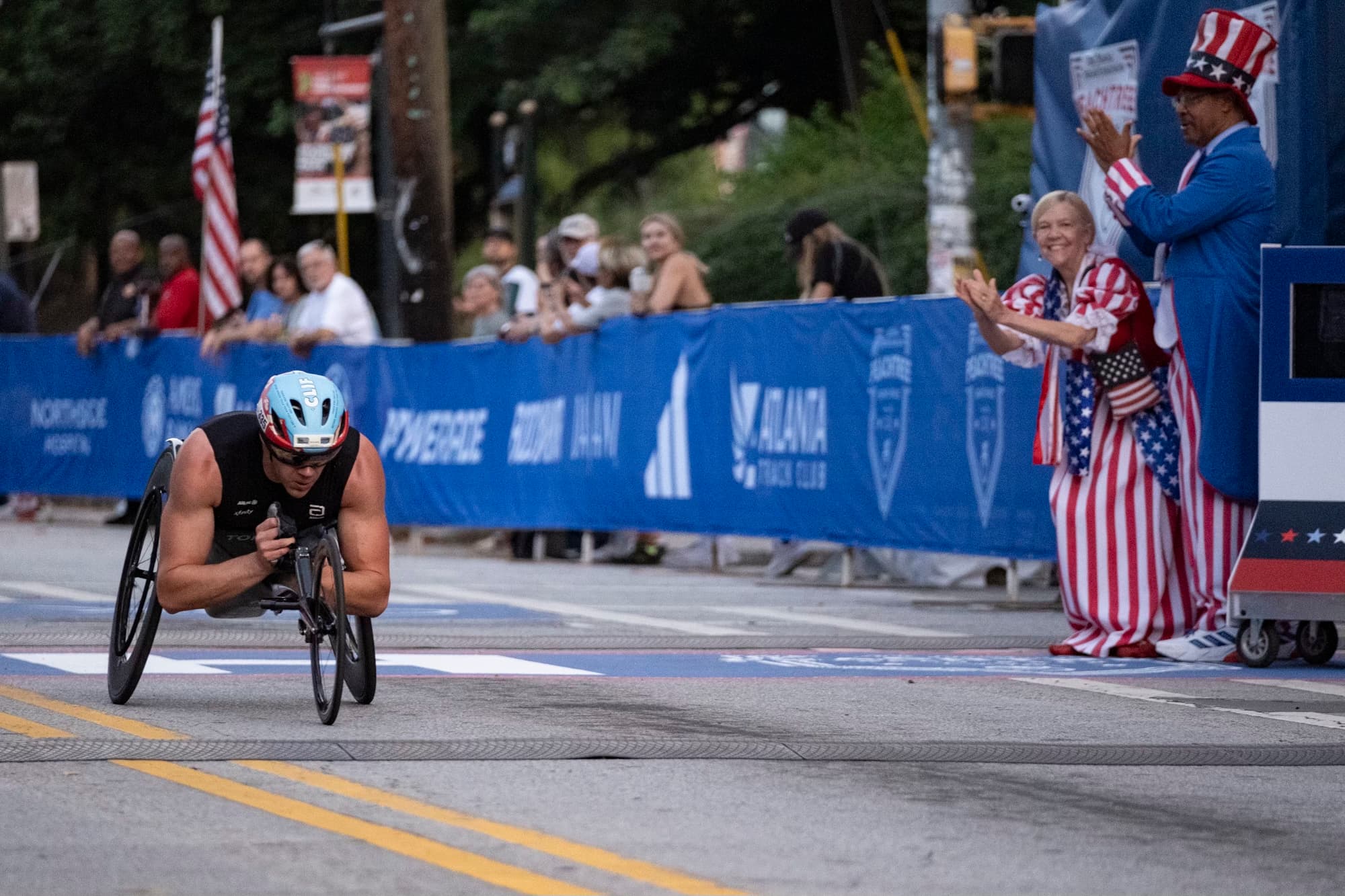 An athlete in a wheelchair crosses the finish line of a race. The lane is lined with American flags and blue barriers displaying sponsor logos. Spectators stand on the side, watching and cheering. The athlete wears a helmet and sports attire.