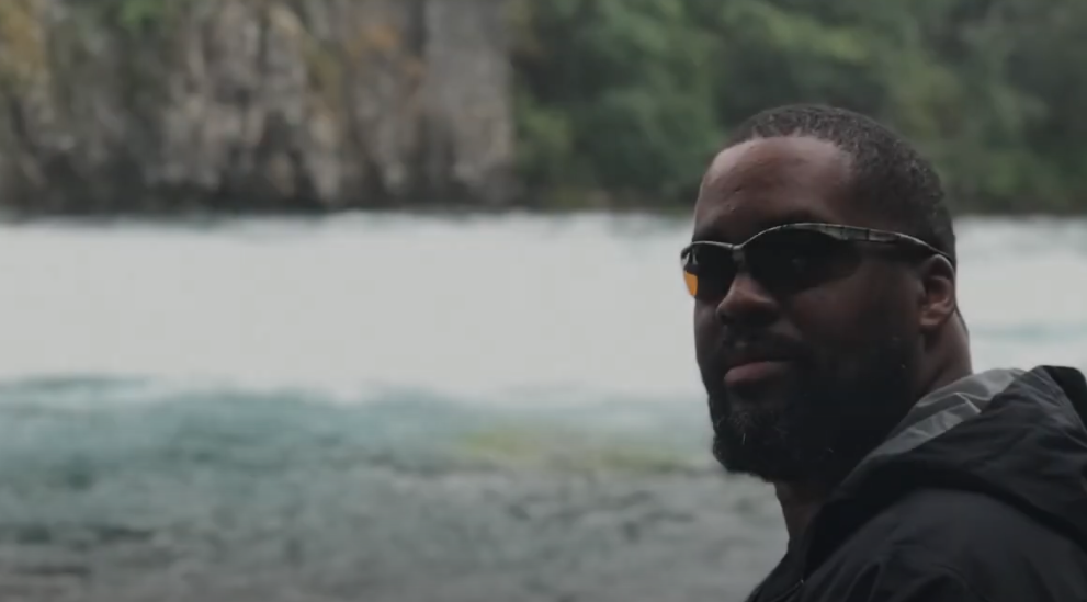 A man wearing sunglasses and a dark jacket stands by a body of water. There is a rocky cliff and lush greenery in the background on a cloudy day.