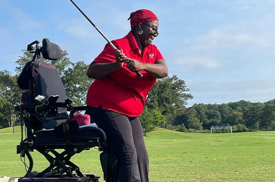 A person wearing a red shirt and dark pants is swinging a golf club on a green golf course. They are positioned beside an adaptive golf chair. Trees and a soccer goal are visible in the background under a blue sky.