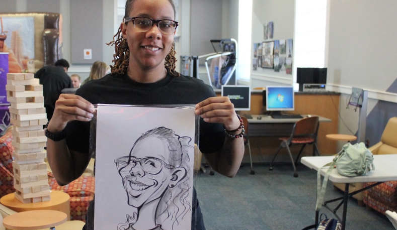 A person with glasses, wearing a black shirt, holds a caricature drawing of themselves, smiling. In the background, there are computers, chairs, a Jenga game, and people in a room with natural light.