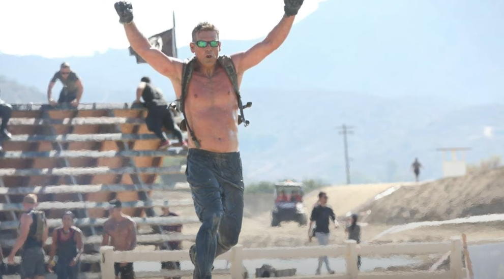 A shirtless man wearing sunglasses and gloves triumphantly raises his arms while running in a muddy outdoor obstacle course. Other participants and a wooden structure are visible in the background under clear skies.