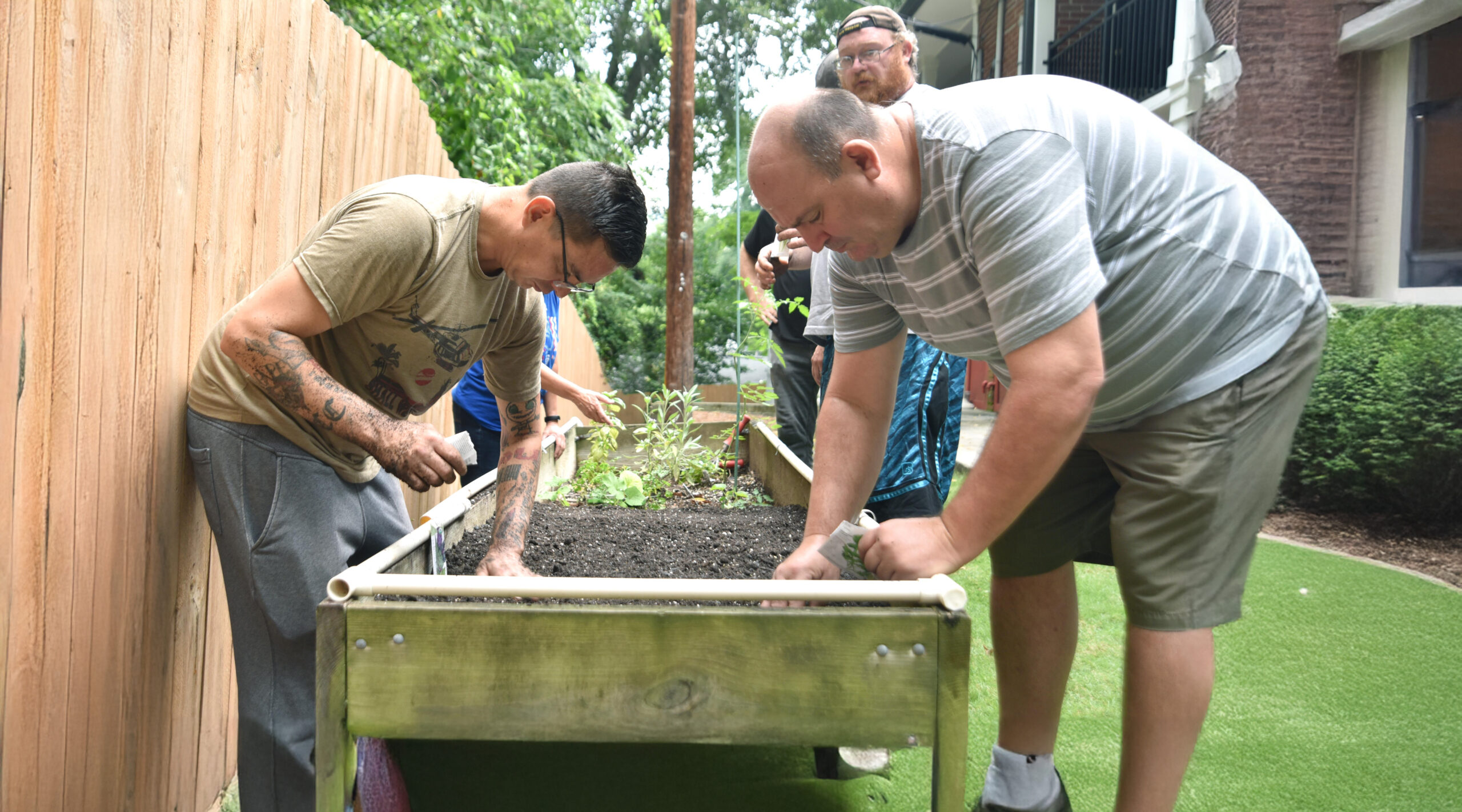 A woman in a green shirt helps two men with plant arrangements. They are seated at a table with various plants and gardening supplies. One man wears a white shirt, and the other a green shirt and camouflage hat, all appearing focused on the task.