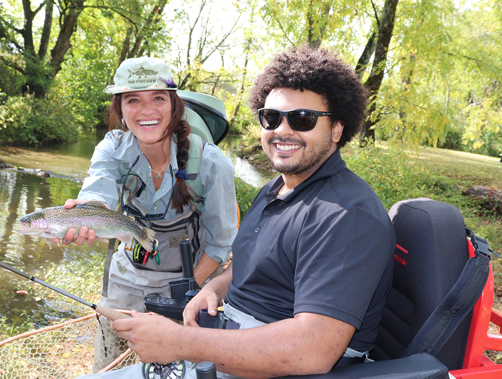 Two people smiling near a riverbank. The person on the left, wearing outdoor gear and a cap, holds a fish. The person on the right, wearing sunglasses and seated in a wheelchair, holds a fishing rod. Trees and a calm river are in the background.