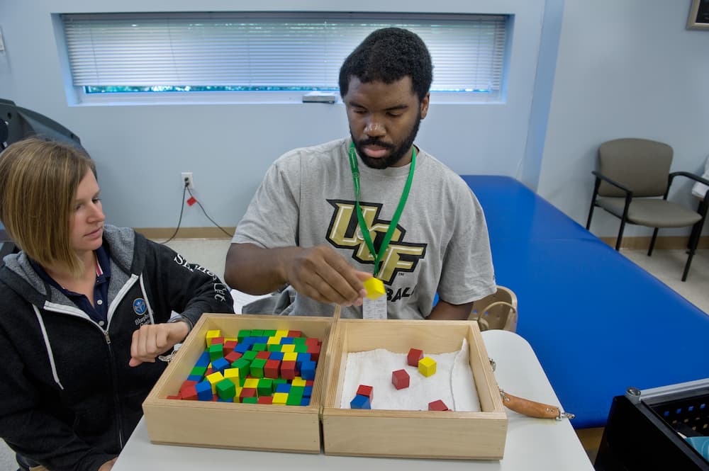 A man in a gray T-shirt sits at a table, picking up a colored block from a box filled with various blocks, as part of an activity. A woman sits beside him, observing his progress.