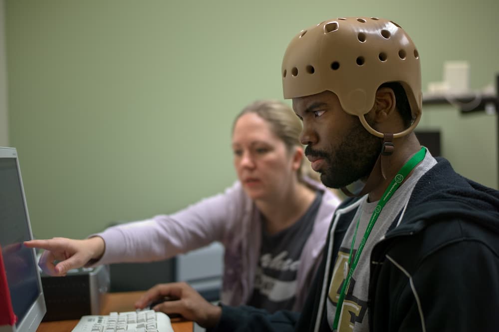 A man wearing a protective helmet works on a computer, while a woman from Shepherd Center beside him points at the screen, offering guidance. They are focused and seated in a room with a green wall.