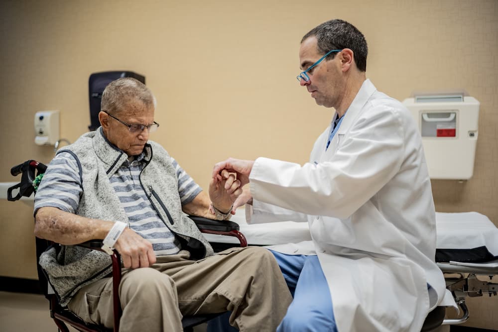 A doctor in a white coat examines an elderly man's wrist in a medical office. The elderly man is sitting in a wheelchair, wearing glasses and a striped shirt with a vest, looking down at his hand.