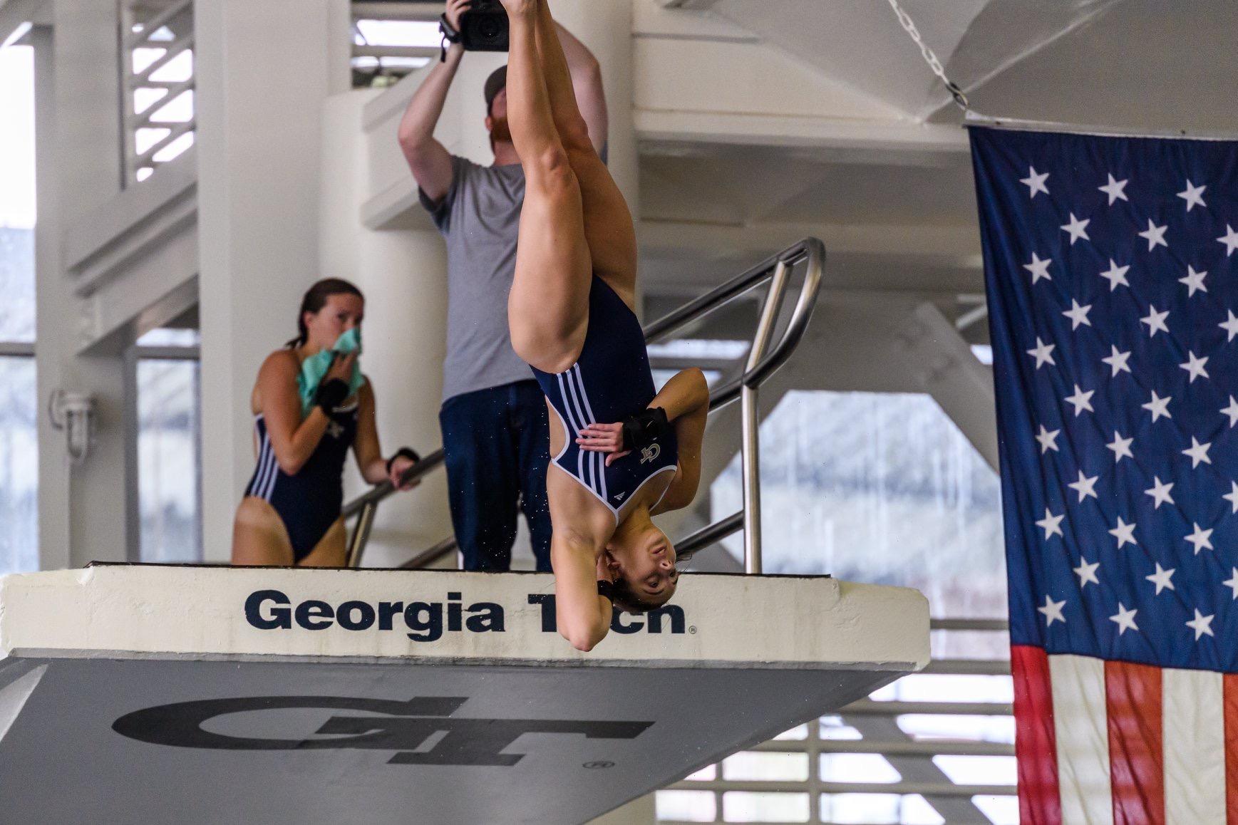 A diver in a navy swimsuit is upside down, diving off a platform at Georgia Tech. Another person in a swimsuit and a person with a camera stand nearby. An American flag hangs in the background.