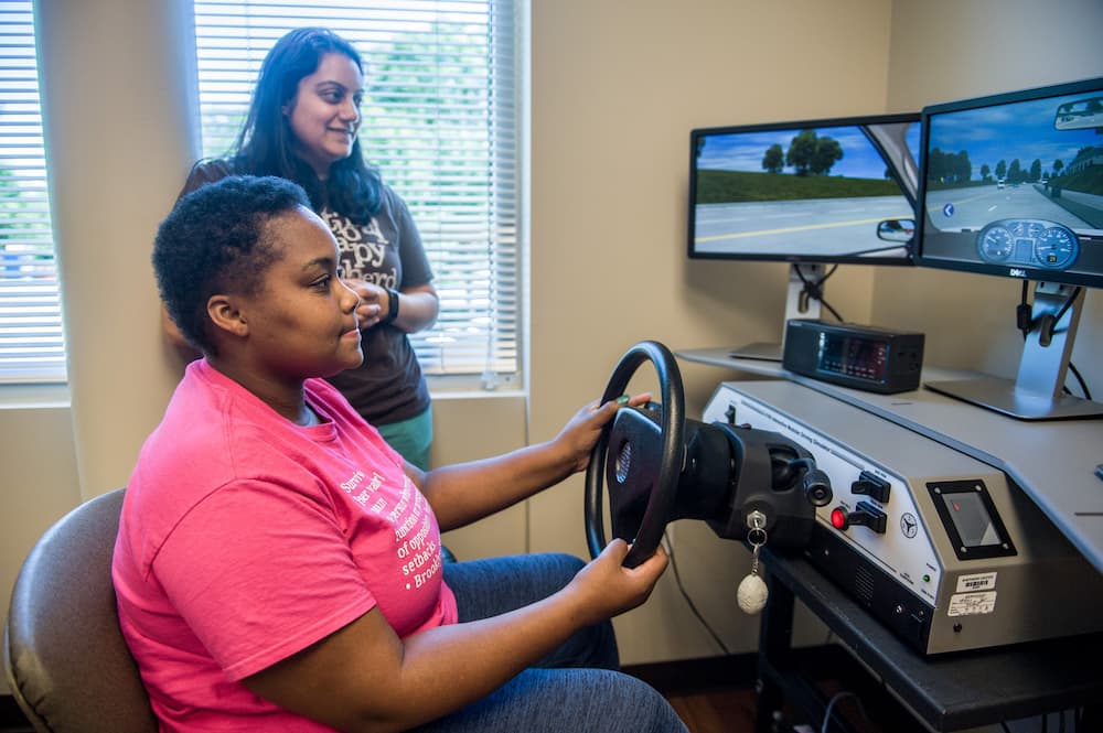 A person sits at a driving simulator in a room, holding the steering wheel and focusing on the screen showing a road. Another person stands nearby, observing. There are three monitors displaying the simulation environment.